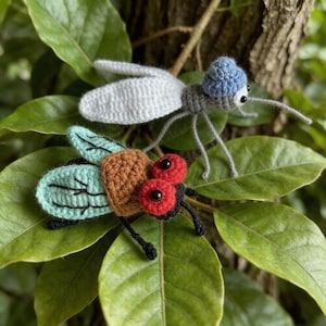 May include: Two handmade crochet insects: a fly and a mosquito. The fly has a brown body, red eyes, and turquoise wings. The mosquito has a grey body, white wings, and a blue hat. Both are set against a backdrop of green leaves.