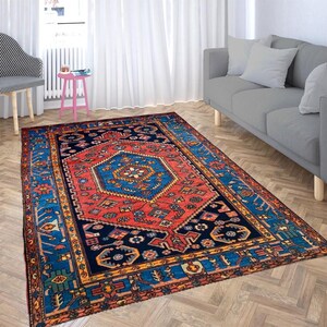 May include: A rectangular area rug with a central red and blue geometric design. The rug has a navy blue border with intricate patterns and is placed on a hardwood floor. The rug's design includes shades of red, blue, and gold.