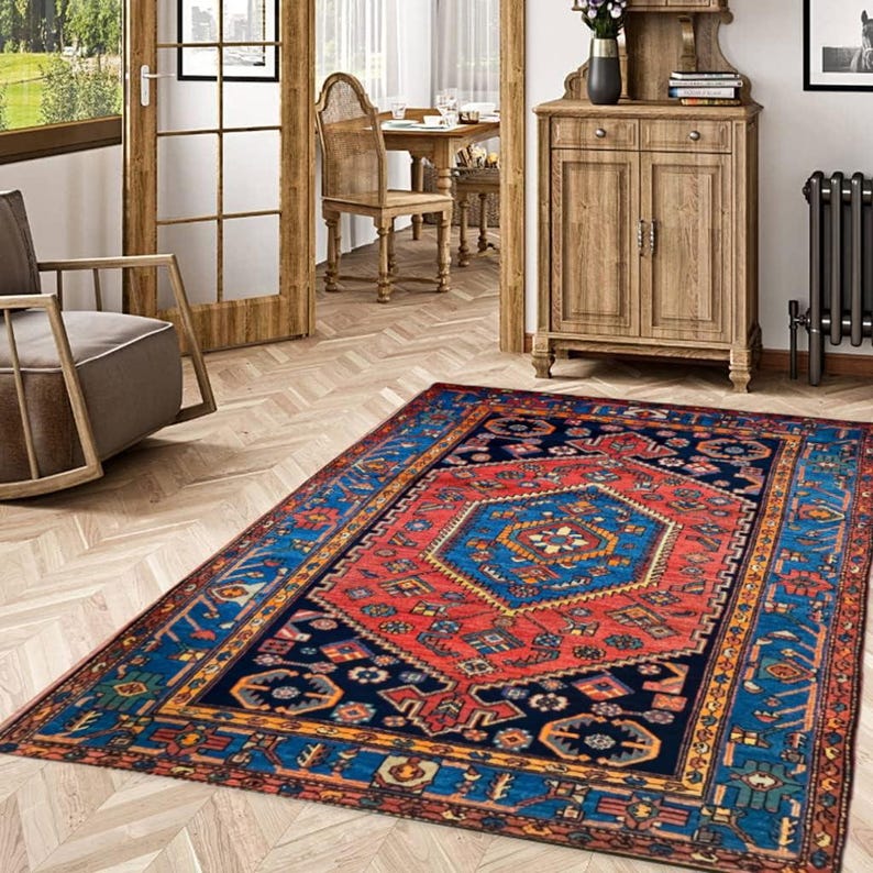 May include: A rectangular area rug with a central geometric design in red and blue. The rug has a dark blue border with intricate patterns and a red central field. The rug is placed on a light-colored wooden floor.