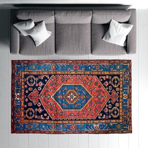 May include: An overhead view of a gray sofa with white pillows and a vibrant, rectangular rug. The rug features a central geometric design in red, blue, and gold, with intricate border patterns. The rug is placed on a white floor.