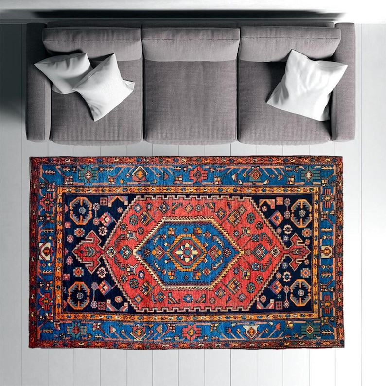 May include: An overhead view of a gray sofa with white pillows and a vibrant, rectangular rug. The rug features a central geometric design in red, blue, and gold, with intricate border patterns. The rug is placed on a white floor.