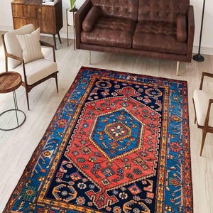 May include: A rectangular area rug with a central geometric design in red and blue, surrounded by a navy blue border with intricate patterns. The rug is placed in a living room setting with brown leather furniture and wooden accents.