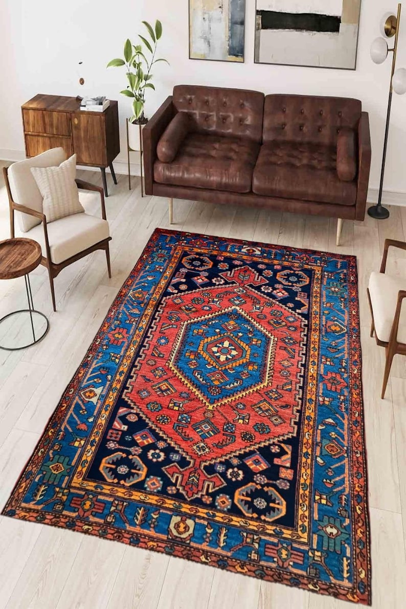 May include: A rectangular area rug with a central geometric design in red and blue, surrounded by a navy blue border with intricate patterns. The rug is placed in a living room setting with brown leather furniture and wooden accents.