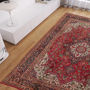 May include: A red and cream-colored Persian rug with an intricate floral pattern dominates the scene. A white robotic vacuum cleaner sits on the wooden floor near the rug. A white lamp and cabinet are in the background.