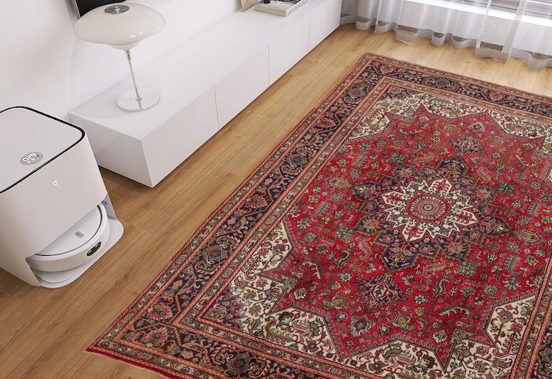 May include: A red and cream-colored Persian rug with an intricate floral pattern dominates the scene. A white robotic vacuum cleaner sits on the wooden floor near the rug. A white lamp and cabinet are in the background.
