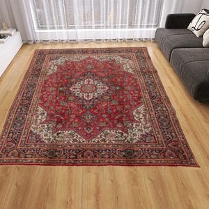 May include: A rectangular, ornate red rug with a central medallion design and intricate border details. The rug is placed on a wooden floor in a living room setting, with a sofa and other furniture visible in the background.