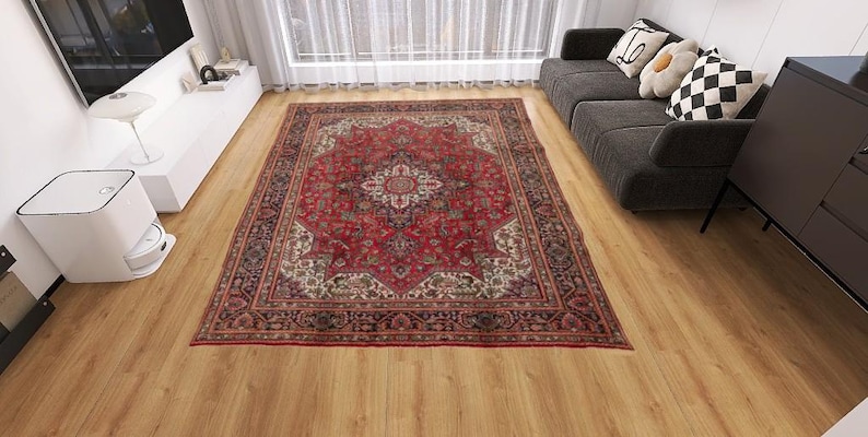 May include: A rectangular, ornate red rug with a central medallion design and intricate border details. The rug is placed on a wooden floor in a living room setting, with a sofa and other furniture visible in the background.