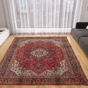 May include: A large, rectangular red Persian rug with an intricate floral pattern dominates a living room. The rug's central field is a deep red, surrounded by a detailed border. The room features a gray sofa, wooden floors, and a large window.