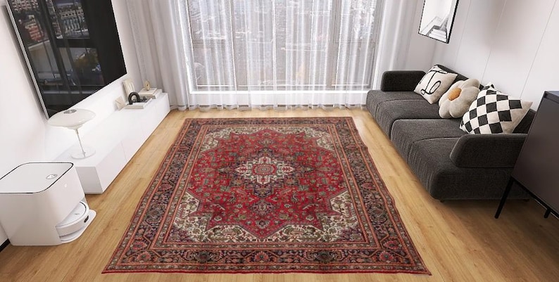 May include: A large, rectangular red Persian rug with an intricate floral pattern dominates a living room. The rug's central field is a deep red, surrounded by a detailed border. The room features a gray sofa, wooden floors, and a large window.