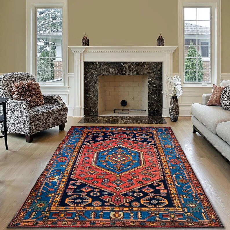 May include: A rectangular, patterned rug with a central geometric design in red and blue. The rug features a navy blue border with intricate patterns and is placed on a hardwood floor in a living room setting.