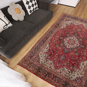 May include: A red and ivory oriental rug with a central medallion design and intricate border. The rug is placed on a hardwood floor next to a dark gray sofa with decorative pillows. The rug's dimensions are not visible.