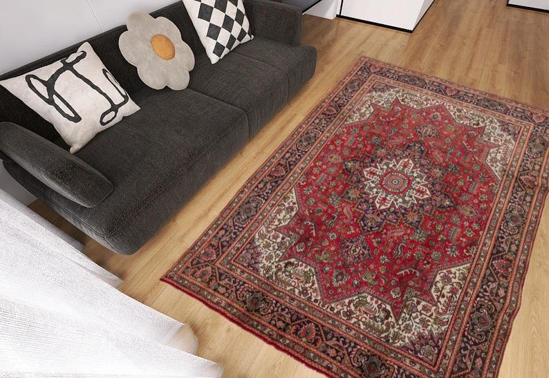 May include: A red and ivory oriental rug with a central medallion design and intricate border. The rug is placed on a hardwood floor next to a dark gray sofa with decorative pillows. The rug's dimensions are not visible.