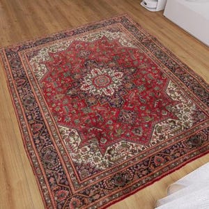 May include: A rectangular red oriental rug with an intricate floral pattern. The rug features a central medallion design with a border of repeating patterns in shades of red, cream, and brown. The rug is placed on a wooden floor.
