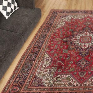 May include: A large, rectangular red rug with an intricate, geometric pattern. The rug features a central medallion design and a border with floral and geometric motifs. The rug is placed on a wooden floor next to a dark gray sofa with decorative pillows.