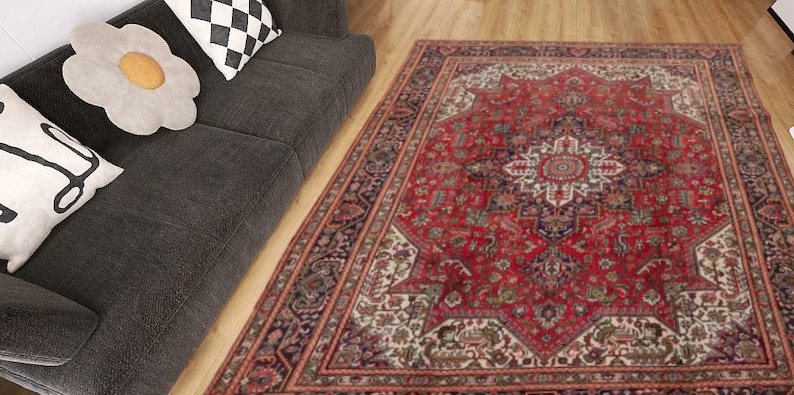 May include: A large, rectangular red rug with an intricate, geometric pattern. The rug features a central medallion design and a border with floral and geometric motifs. The rug is placed on a wooden floor next to a dark gray sofa with decorative pillows.