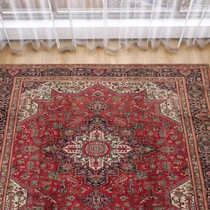 May include: A rectangular, red Persian rug with an intricate, symmetrical design. The rug features a central medallion and detailed borders in shades of red, cream, black, and green. It is placed on a wooden floor near a window and a dark gray sofa.