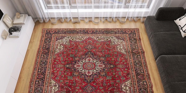 May include: A rectangular, red Persian rug with an intricate, symmetrical design. The rug features a central medallion and detailed borders in shades of red, cream, black, and green. It is placed on a wooden floor near a window and a dark gray sofa.