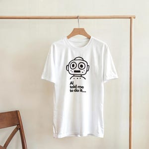 Puede incluir: Camiseta blanca con un gráfico de robot negro y el texto "AI told me to do it...". El diseño del robot presenta una cabeza circular, dos ojos grandes y una boca rectangular. La camiseta está colgada en una percha de madera.
