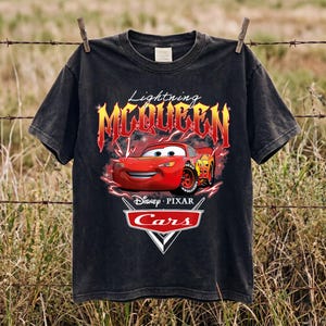 May include: A black short-sleeve t-shirt featuring a graphic of the red Lightning McQueen character from the Disney Pixar movie Cars. The design includes the text Lightning MCQUEEN in a fiery font and the official Cars logo with Disney Pixar text.