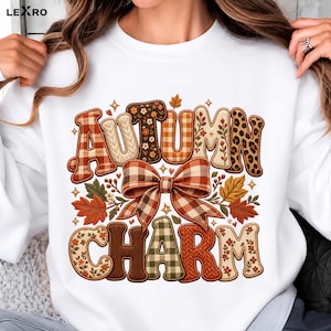 May include: A white sweatshirt featuring the text AUTUMN CHARM in large, patterned letters. The letters include plaid, floral, cable knit, and leopard print designs. A large brown and white plaid bow sits between the two words.