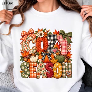 Op de afbeelding: Een wit sweatshirt met ronde hals en een kleurrijke afbeelding met de tekst COZY SEASON. Het ontwerp bevat pompoenen, herfstbladeren, dennenappels en geruite strikken, met letters versierd in bloemen-, gingham- en kabelbreipatronen.