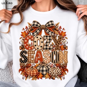 Peut inclure: Un sweat-shirt blanc à manches longues avec une impression graphique portant le texte COZY SEASON. Le motif comprend des citrouilles, des feuilles d'automne, des glands et des nœuds aux motifs variés comme le tartan, le léopard et les pois.