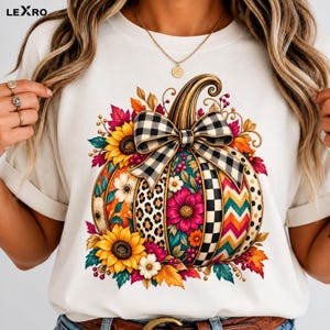 Peut inclure: Un t-shirt blanc orné d'un grand motif de citrouille décorée de tournesols, de feuilles d'automne et de divers motifs incluant des taches de léopard, un damier noir et blanc et des chevrons, surmontée d'un nœud à carreaux vichy.