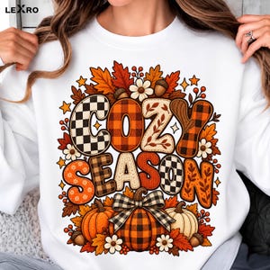 Peut inclure: Un sweat-shirt blanc à manches longues avec une impression graphique portant le texte COZY SEASON. Le motif comprend des citrouilles orange, noires et blanches, des feuilles d'automne, des glands et des fleurs, avec des lettres à motifs.