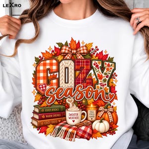 Può includere: Una felpa bianca con una stampa grafica del testo COZY season. Il design include un fiocco a quadri, foglie autunnali, una pila di tre libri etichettati GOOD BOOKS, WARM DRINKS, COZY DAYS, una tazza con SWEATER WEATHER, una candela e delle zucche.