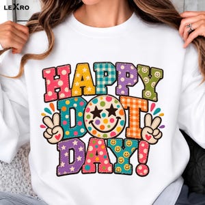 Könnte beinhalten: Ein weißes Langarm-Sweatshirt mit einer bunten Grafik und dem Schriftzug HAPPY DOT DAY. Die Buchstaben sind mit verschiedenen Mustern wie Punkten, Sternen, Karos und Smileys gefüllt. Zwei Hände zeigen das Friedenszeichen neben dem Text.