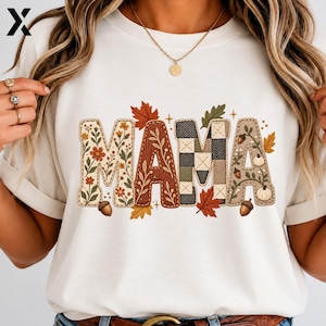 May include: A white short-sleeved t-shirt featuring the word MAMA in large, decorative letters. Each letter has a unique pattern including floral prints, ribbed textures, and checkered designs, accented by small illustrations of autumn leaves, acorns, and pumpkins.