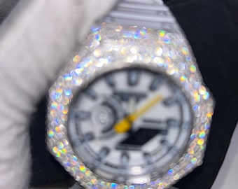 Könnte beinhalten: Eine weiße Uhr mit weißem Zifferblatt und gelben Zeigern. Die Uhr ist mit funkelnden, irisierenden Edelsteinen um die Lünette besetzt. Die Uhr wird von einer behandschuhten Hand gehalten.