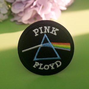 May include: A black circular patch featuring the words "PINK FLOYD" in white, surrounding a blue triangle with a white beam of light and a rainbow. The patch is likely an embroidered design, possibly for clothing or accessories.