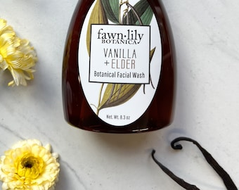 May include: A brown bottle of fawn-lily botanica vanilla + elder botanical facial wash. The bottle has a white label with a botanical illustration of vanilla beans and leaves. The label also includes the text "fawn-lily botanica", "vanilla + elder", and "botanical facial wash". The bottle is 8.3 ounces.
