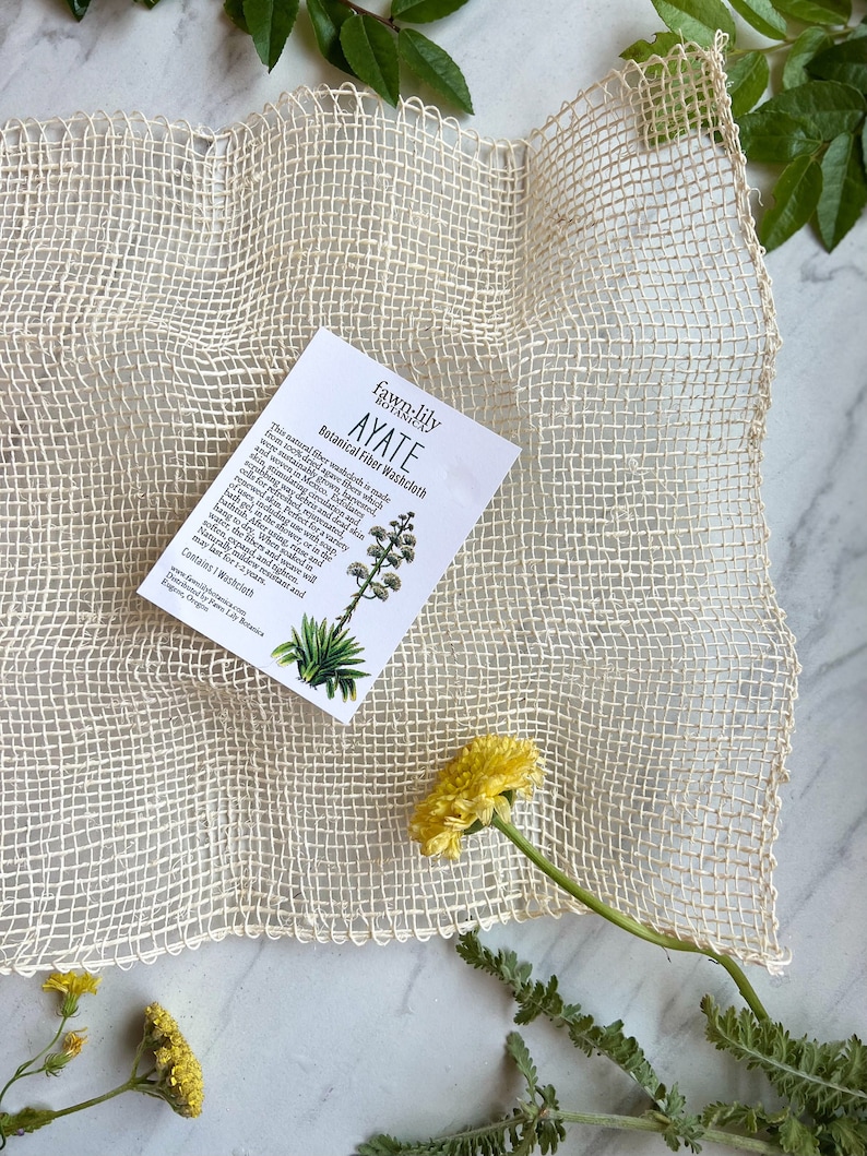 Puede incluir: Un pa&ntilde;o de lavado blanco tejido con fibras de agave, llamado Ayate, se muestra sobre una superficie blanca con una peque&ntilde;a tarjeta que describe el producto. La tarjeta dice "fawn:lily botanicals AYATE Botanical Fiber Washcloth".