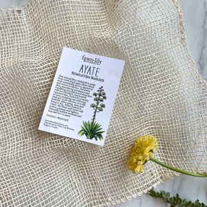 Puede incluir: Un pa&ntilde;o de lavado blanco hecho de fibras de agave, llamado Ayate, se muestra sobre un fondo de fibra natural. El pa&ntilde;o de lavado se fabrica en M&eacute;xico y se cosecha de forma sostenible. El texto de la tarjeta dice "fawn-lily botanica Ayate Botanical Fiber Washcloth".