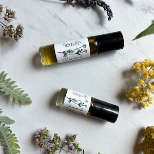 May include: Two small glass bottles with a roll-on applicator. The bottles are filled with a clear liquid and have white labels with the text "fawn-lily botanica" and "Chamomile." The bottles are surrounded by various flowers and greenery.