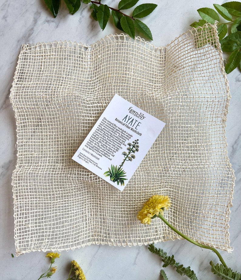 Puede incluir: Una toallita de lavado de malla blanca tejida a partir de fibras naturales. La toallita est&aacute; sobre una superficie blanca con una peque&ntilde;a tarjeta que dice "Fawn-Lily Botanicals AYATE Botanical Fiber Washcloth".