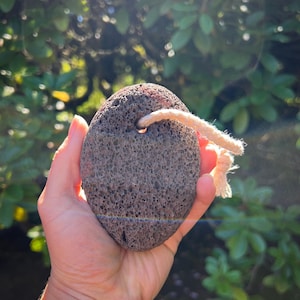 Puede incluir: Una piedra pómez gris oscuro con una superficie texturizada y una cuerda de color crema adjunta. La piedra tiene forma ovalada y parece porosa. El fondo está borroso con follaje verde.