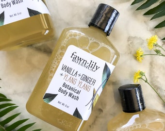 May include: Three clear plastic bottles of body wash with a yellow liquid. The bottles are labeled "fawn-lily BOTANICA" and "VANILLA+GINGER + YLANG YLANG Botanical Body Wash". The bottles are on a white marble surface with green fern leaves and yellow flowers.