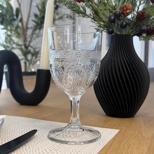 May include: A clear, patterned glass goblet with a decorative rim, set on a table with a neutral-toned placemat. A black vase with a spiral design holds greenery and berries. A black candle and a decorative black object are also visible.