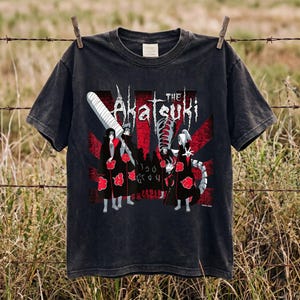 May include: A black short-sleeve t-shirt featuring a graphic print of anime characters in black cloaks with red clouds. The text "THE Akatsuki" is printed above the characters, set against a background of red and black sunburst rays.