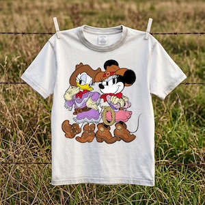 以下が含まれることがあります： クリーム色の半袖Tシャツ。ウエスタンスタイルのカウガール衣装を着たデイジーダックとミニーマウスのグラフィックプリントが施されています。両キャラクターとも茶色のカウボーイハット、赤いネッカチーフ、花柄のディテールが施された茶色のブーツを着用しています。