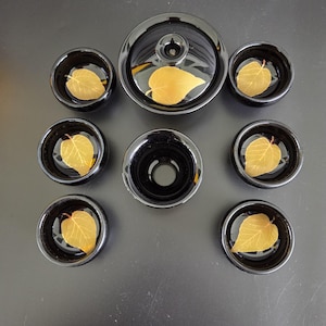 May include: A black ceramic tea set consisting of a lidded gaiwan and six small tea cups. Each piece features a single, realistic golden-yellow leaf embedded at the bottom of the interior. The set is arranged symmetrically on a dark surface.