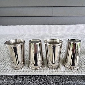 May include: Four silver-colored metal tumblers with a reflective surface. The tumblers are cylindrical with a slightly flared rim. They are arranged in a row on a white, textured surface, reflecting the surrounding environment.