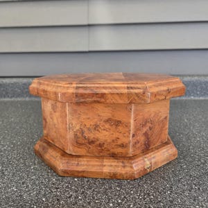 May include: An octagonal wooden box featuring a rich, swirling burl wood grain pattern. The container has a beveled lid and a matching wide base, showcasing a polished, warm brown finish with natural wood textures throughout the entire structure.