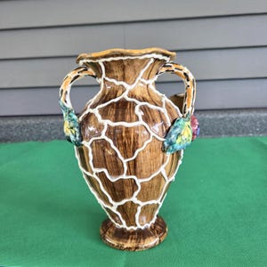 May include: A ceramic vase with a brown wood-grain finish and a raised white lattice pattern overlay. The vase features two side handles, each adorned with sculpted, multi-colored leaves and fruit accents at the base of the handles.