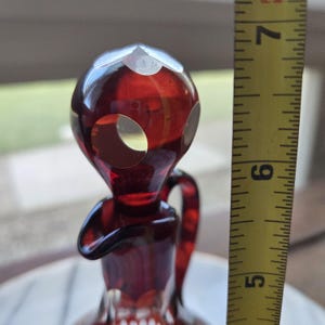 May include: A ruby red glass decanter with a unique alien-like design. The decanter has a round head with three circular cutouts and a handle. The height of the decanter is approximately 6 inches.