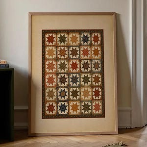 May include: A framed rectangular wall art print featuring a repeating grid of star-shaped quilt patterns in muted tones of rust, olive green, navy blue, and cream. The artwork is enclosed in a light-colored wooden frame with a wide, plain border.