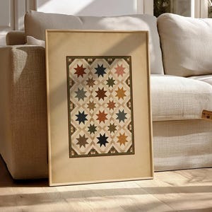 May include: Framed artwork featuring a star quilt design in a variety of colors, including red, blue, and green. The artwork is displayed in a light-colored wooden frame and leans against a beige sofa in a well-lit room. The artwork adds a touch of rustic charm.